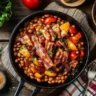 Overhead shot of baked beans in a skillet, topped with crispy bacon, caramelized onions, and bell peppers, surrounded by fresh ingredients.